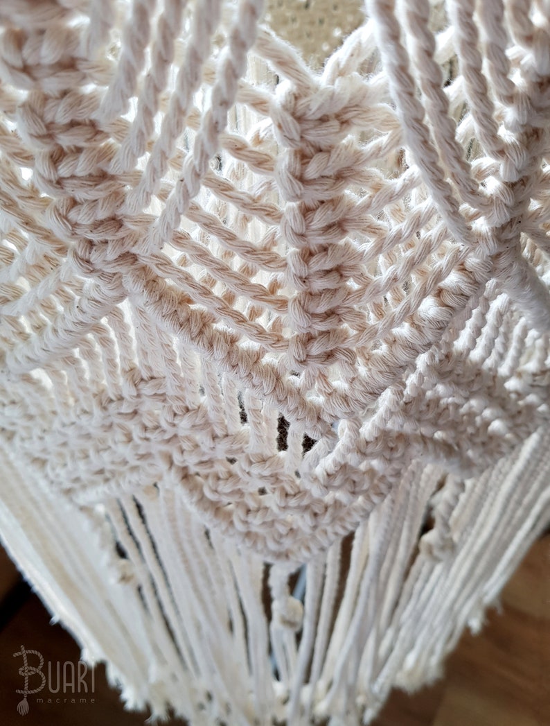 May include: A close-up of a white macrame wall hanging with a geometric pattern and fringe. The macrame is made with natural cotton cord.