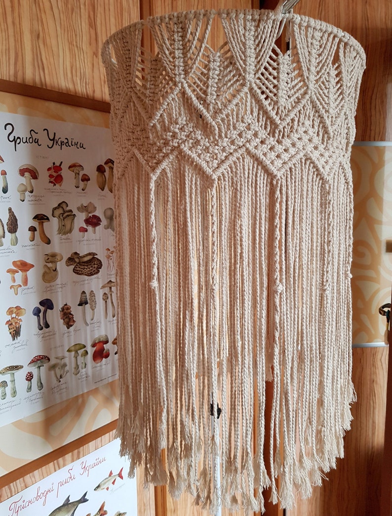 May include: A beige macrame lampshade with a geometric pattern and long fringe. The lampshade is hanging from a ceiling fixture.