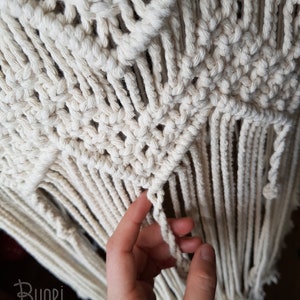 May include: Close-up of a white macrame wall hanging with a hand holding a fringe. The macrame is made with a knot pattern that creates a geometric design. The fringe is long and flowing.