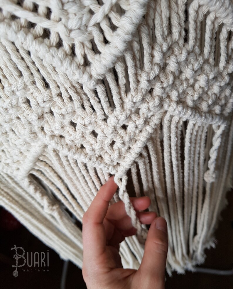 May include: Close-up of a white macrame wall hanging with a hand holding a fringe. The macrame is made with a knot pattern that creates a geometric design. The fringe is long and flowing.