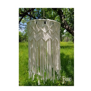 May include: A white macrame hanging lamp with a geometric pattern and long fringe. The lamp is hanging from a tree branch in a green grassy field.