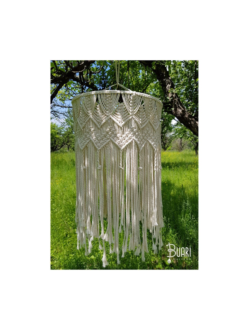 May include: A white macrame hanging lamp with a geometric pattern and long fringe. The lamp is hanging from a tree branch in a green grassy field.