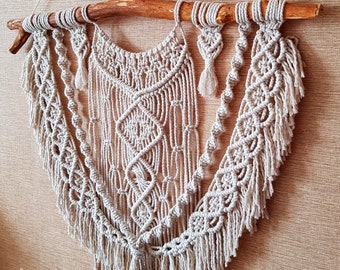 May include: A beige macrame wall hanging with a geometric design, suspended from a natural wooden branch. The handcrafted piece features intricate knotting patterns, tassels, and a textured, bohemian aesthetic. The wall decor is a neutral color.