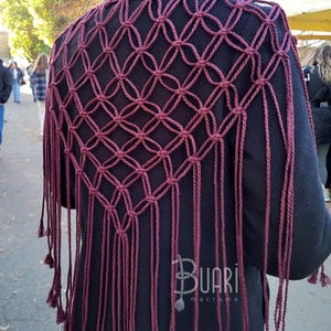 May include: A burgundy macrame shawl with fringe. The shawl is made with a knotting technique that creates a diamond pattern. The shawl is worn over a black jacket.