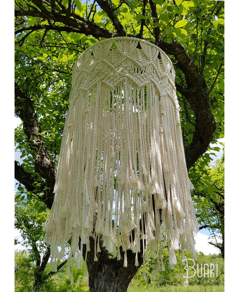 May include: A large, white macrame hanging with a fringe border. The hanging is made of natural cotton cord and is suspended from a tree branch.