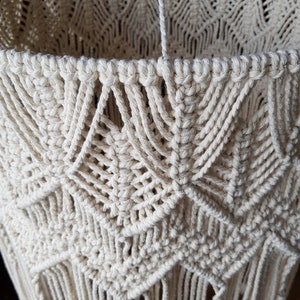May include: A close-up of a cream-colored macrame lampshade with a geometric pattern and fringe. The lampshade is made of natural cotton cord and has a delicate, bohemian style.