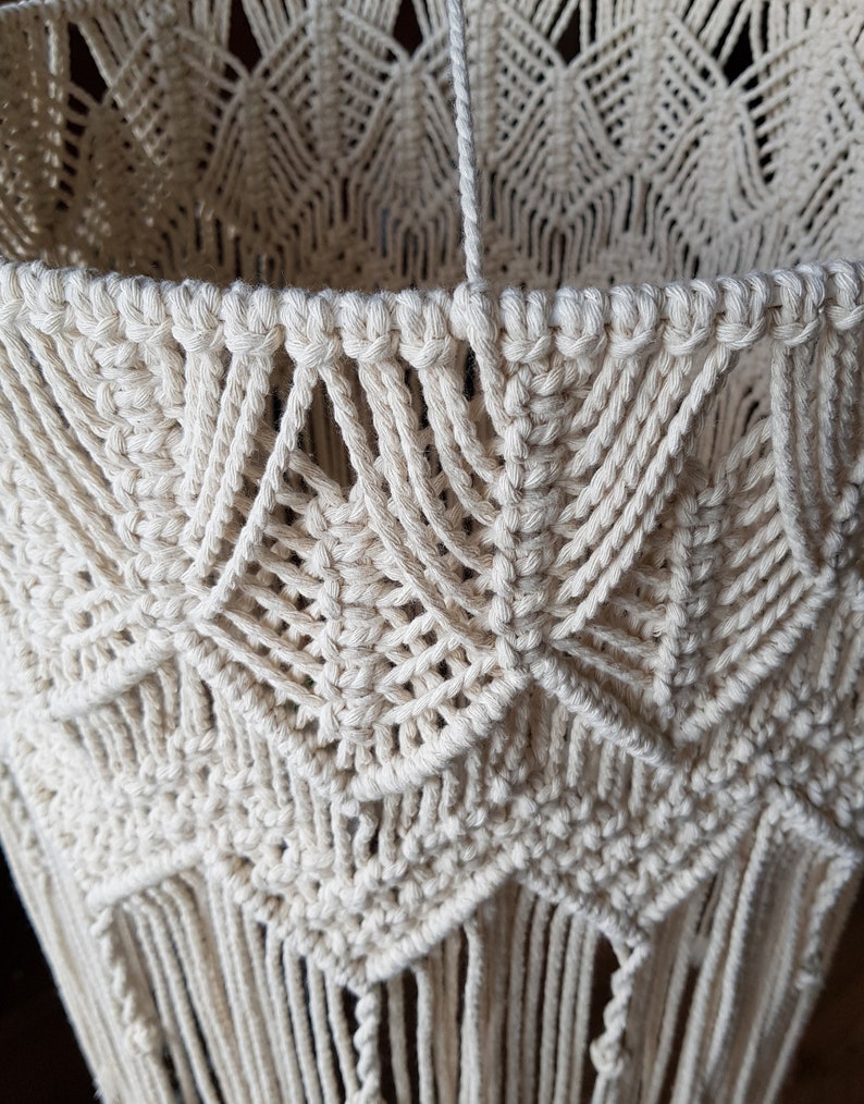 May include: A close-up of a cream-colored macrame lampshade with a geometric pattern and fringe. The lampshade is made of natural cotton cord and has a delicate, bohemian style.
