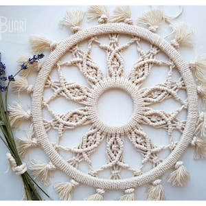 Puede incluir: Un gran tapiz de macramé de color crema con un diseño circular y detalles de flecos. El macramé está hecho con un cordón grueso de fibra natural y tiene un patrón delicado e intrincado.