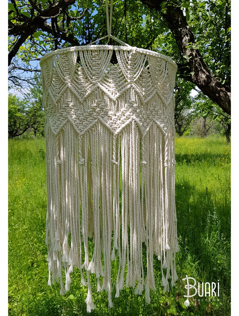May include: A white macrame lampshade hanging from a tree branch. The lampshade is made of a thick, natural fiber rope and has a geometric pattern. The lampshade is hanging in a garden setting with green grass and trees in the background.