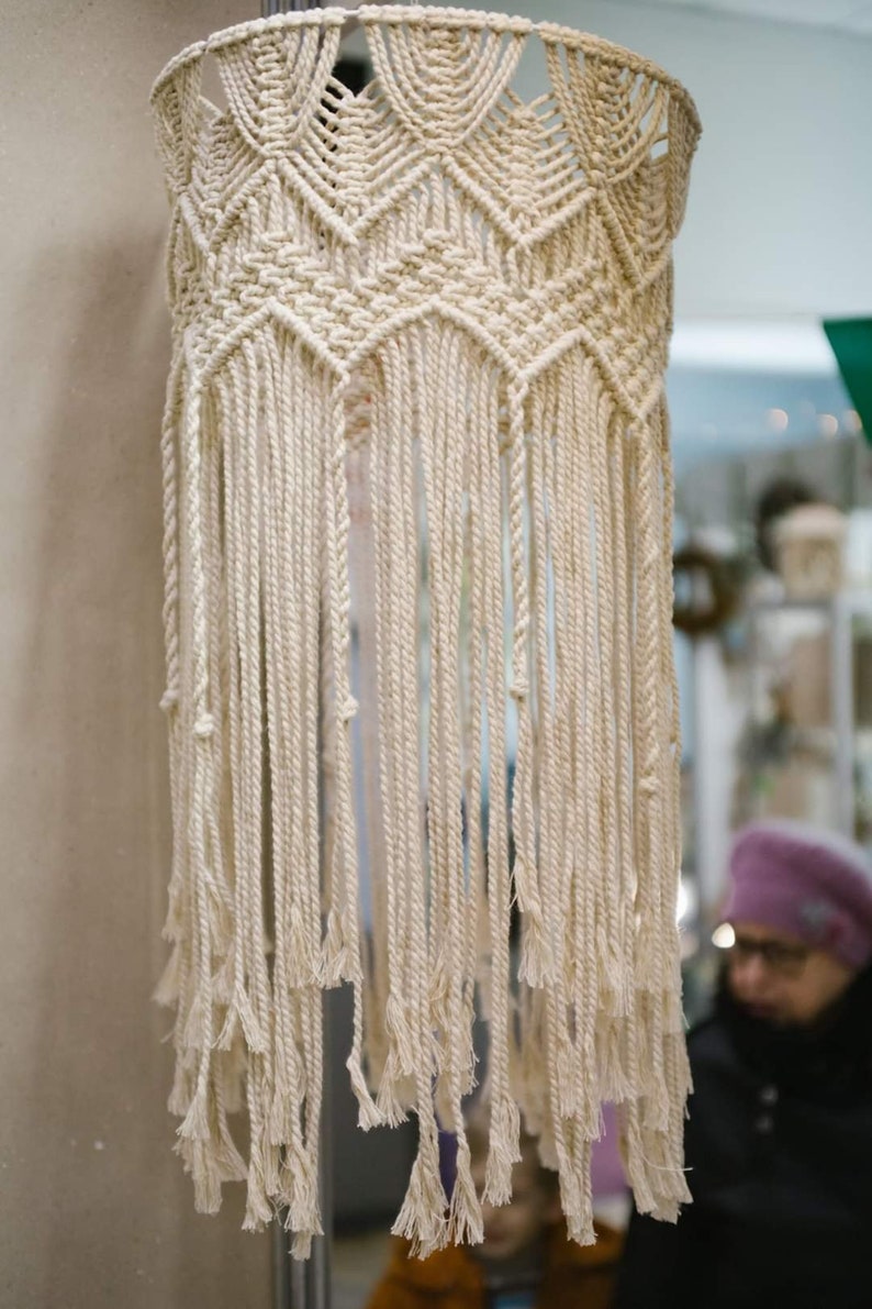 May include: A white macrame hanging lampshade with a geometric pattern and long fringe.