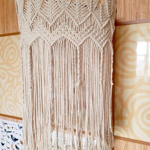 May include: A white macrame wall hanging with a geometric pattern and long fringe. The hanging is made of cotton cord and is attached to a metal rod.