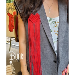 May include: A red heart-shaped macrame piece with long fringe hangs from a gray blazer. The macrame is made with red cord and has a heart shape at the top. The blazer is a classic style with a button closure.