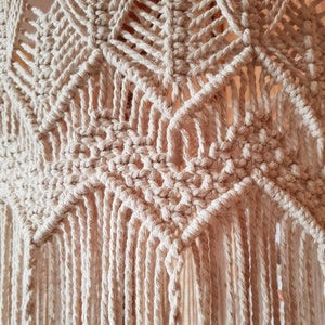 May include: Close-up of a beige macrame wall hanging with a geometric pattern and fringe. The macrame is made with natural cotton cord.