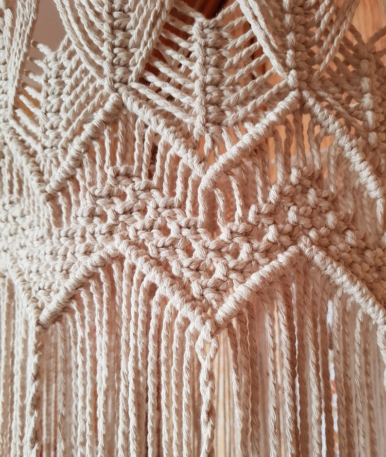 May include: Close-up of a beige macrame wall hanging with a geometric pattern and fringe. The macrame is made with natural cotton cord.