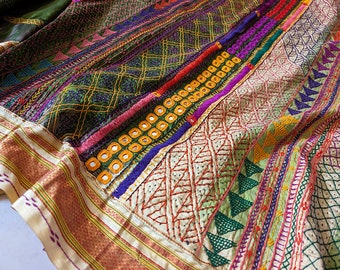 May include: Close-up of a richly embroidered textile with a variety of patterns and colors. The fabric features intricate stitching in shades of green, purple, orange, and yellow, with mirrored accents. The border has a gold and cream striped design.