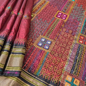 以下が含まれることがあります： 深紅、赤、金色の豊かな刺繍が施された織物。カラフルな糸と小さな鏡のアクセントで複雑な模様が特徴です。縁には黒と金の幾何学模様があります。この織物は、装飾品または衣服の一部である可能性があります。