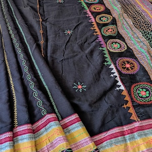 Può includere: Un tessuto nero con intricati ricami multicolori. Il design include motivi floreali circolari, motivi geometrici a zigzag e cuciture lineari in tonalità vivaci di verde, arancione, rosa e giallo lungo il bordo.