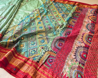 May include: A detailed textile with intricate embroidery in shades of green, blue, yellow, and red. The design features geometric patterns and floral motifs, with a red border. The fabric appears to be soft and flowing.