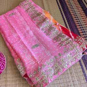 May include: A pink textile with intricate gold embroidery and a patterned design. The fabric has a delicate texture and is folded, revealing a glimpse of orange and red accents. The overall aesthetic is ornate and decorative.