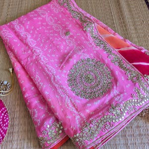 May include: A vibrant pink saree with intricate white dot patterns and gold embellishments. The saree features a detailed border and a large circular design. A matching pink pouch with a pearl necklace is visible.