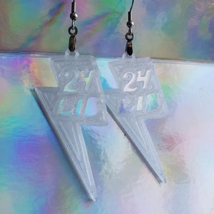 Puede incluir: Un par de pendientes transparentes en forma de rayo. Cada pendiente presenta el número "24" y las letras "LIB" grabadas en el diseño. Cuelgan de ganchos plateados. El fondo es una superficie holográfica iridiscente.