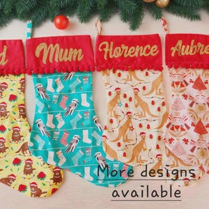 May include: Four Christmas stockings, each with a name: "Dad", "Mum", "Florence", and "Aubrey". They have red tops with gold lettering and pom-pom trim. The patterned bodies display festive designs with animals and Christmas trees. The text "More designs available" is visible.