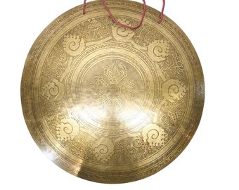 May include: A large, round, gold-colored gong with intricate designs and a red rope handle. The gong has a raised center with a circular design and is surrounded by a series of concentric circles.