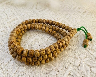 May include: A strand of prayer beads made of light brown, speckled seeds. The beads are strung together with a green cord, forming a loop. The beads are arranged in a double strand.
