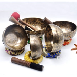 May include: A selection of bronze singing bowls with wooden mallets. The bowls are of varying sizes and rest on colourful cushions. Some bowls feature etched designs and text. The mallets have red and black felt striking surfaces.