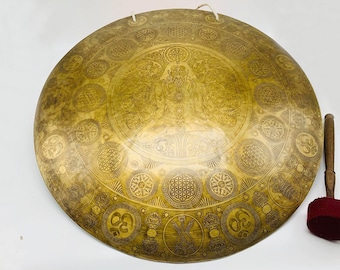 May include: A large, round, gold-colored gong with intricate designs and symbols. The gong is hanging from a string and has a wooden mallet with a red felt head.