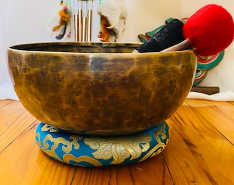 May include: A large, antique-looking, brass singing bowl with a blue and gold patterned cushion. A red felt mallet is resting on the bowl.