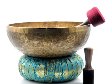 May include: A gold singing bowl with intricate carvings of the Om symbol, resting on a blue and gold patterned cushion. A wooden striker with a red felt tip is included.