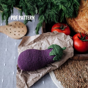 Puede incluir: Un juguete de berenjena de crochet con un cuerpo morado y hojas verdes. El juguete está sobre papel marrón arrugado con tomates rojos y una hogaza de pan en el fondo.