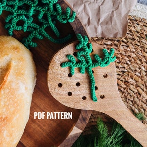 Op de afbeelding: Een houten lepel met een groene gehaakte tak groen erop. De lepel ligt op een houten bord met een brood en meer gehaakt groen. De tekst "PDF PATTERN" staat op het bord.