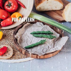 May include: A still-life image featuring fresh produce and handmade crochet items. A wooden bowl holds tomatoes and bell peppers. Three green crochet chili peppers rest on a wooden plate. The text "PDF PATTERN" is visible.