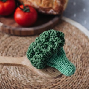 May include: A green crocheted broccoli toy on a wooden spoon. The broccoli is a realistic representation of a real broccoli floret.
