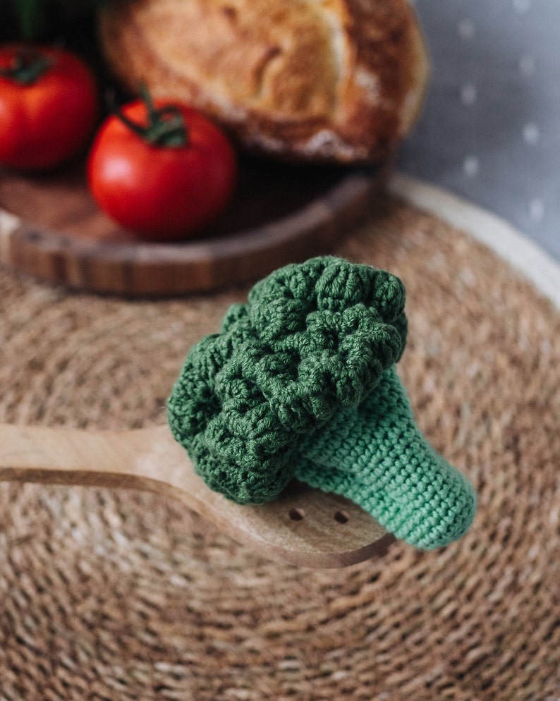 May include: A green crocheted broccoli toy on a wooden spoon. The broccoli is a realistic representation of a real broccoli floret.