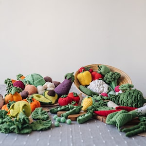 May include: A collection of crocheted vegetables, including a green avocado, red tomato, yellow bell pepper, green broccoli, and a purple eggplant, spilling out of a woven basket onto a gray tablecloth.