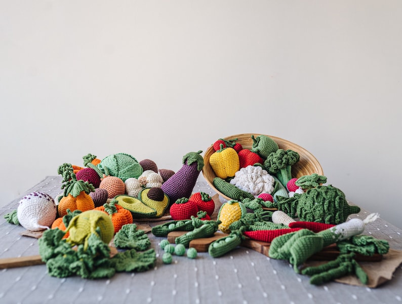 May include: A collection of crocheted vegetables, including a green avocado, red tomato, yellow bell pepper, green broccoli, and a purple eggplant, spilling out of a woven basket onto a gray tablecloth.