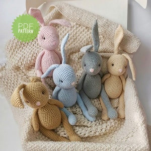 Könnte beinhalten: Fünf gehäkelte Hasen in verschiedenen Farben, darunter rosa, blau, grau und beige, sitzen auf einer weißen Häkeldecke. Die Hasen haben lange Ohren und schauen alle nach vorne. Das Bild enthält den Text "PDF PATTERN".
