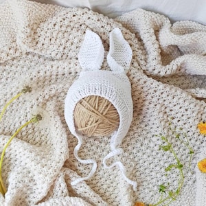 May include: White crocheted bunny hat with long ear flaps and ties. The hat is lying on a white crocheted blanket.