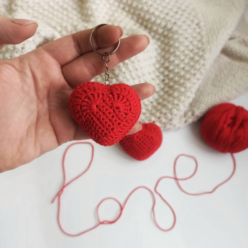 May include: Red crocheted heart keychain with a silver key ring. The heart is hanging from a silver chain. The word "love" is spelled out in red yarn on a white surface.