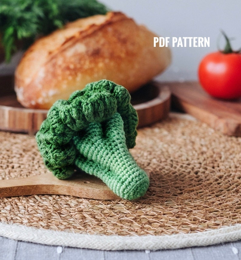 May include: A crocheted green broccoli toy on a woven placemat. The toy is made with green yarn and has a realistic shape. The placemat is brown and has a white border.