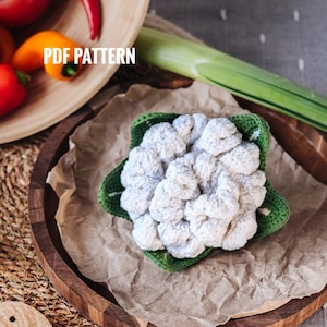Puede incluir: Un coliflor de ganchillo con hojas verdes, sentado sobre una superficie de papel marrón. El coliflor es blanco y tiene una textura realista. La imagen incluye el texto "PDF PATTERN".