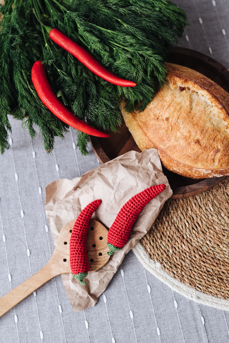 Puede incluir: Dos chiles rojos de ganchillo en una cuchara de madera y papel marr&oacute;n, junto a una hogaza de pan y un manojo de eneldo fresco.
