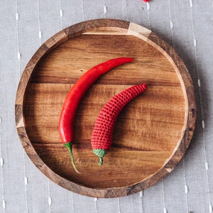 Puede incluir: Tres chiles rojos, dos en un plato de madera y uno en una superficie gris texturizada. Los chiles son de diferentes tama&ntilde;os y formas.
