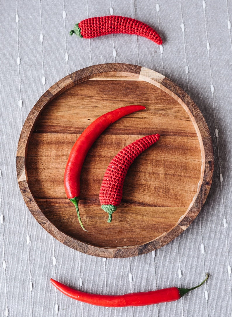 Puede incluir: Tres chiles rojos, dos en un plato de madera y uno en una superficie gris texturizada. Los chiles son de diferentes tama&ntilde;os y formas.