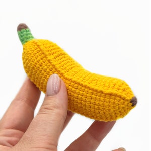 May include: A crocheted banana toy with a yellow body, green stem, and brown tip. The banana is held in a hand.