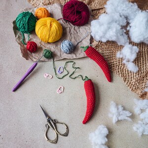 Puede incluir: Primer plano de un proyecto artesanal con ovillos de lana roja, verde, amarilla y azul, dos chiles rojos tejidos a crochet, un par de tijeras y un gancho de crochet. Los ovillos de lana est&aacute;n dispuestos sobre un fondo de papel marr&oacute;n con bolas de algod&oacute;n blancas.