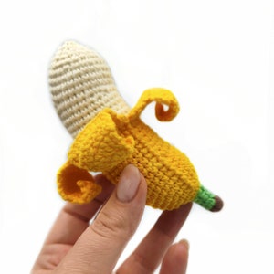 May include: A crocheted banana toy with a yellow peel and a cream-colored top. The banana is held in a hand.
